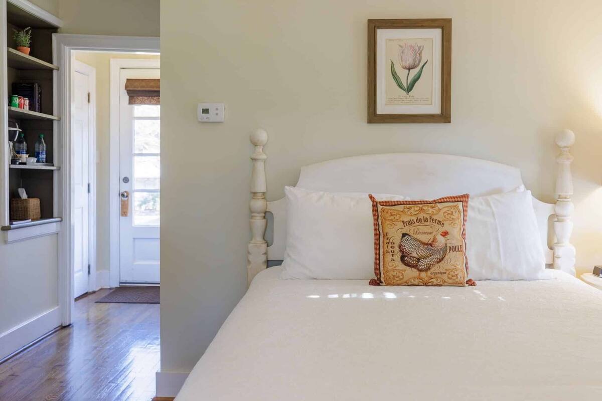 A bedroom with a white bed, a wooden headboard, a framed botanical print on the wall, and a decorative pillow with a rooster design.