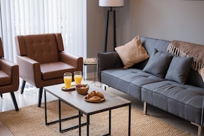 Ein modernes Wohnzimmer mit einem grauen Sofa, einem braunen Sessel und einem Couchtisch mit Frühstücksangeboten.