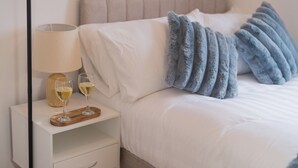 A neatly made bed with white linens and a blue throw blanket, a bedside table with a lamp and two glasses of white wine, and a wooden tray.