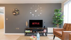 A living room with a wall clock, a flat-screen TV displaying 'NETFLIX', a coffee table with snacks and a water bottle, and a brown leather armchair.