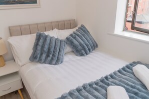 Uma cama bem arrumada com um cobertor macio branco e azul, uma cabeceira de madeira e uma janela com vista para edifícios.