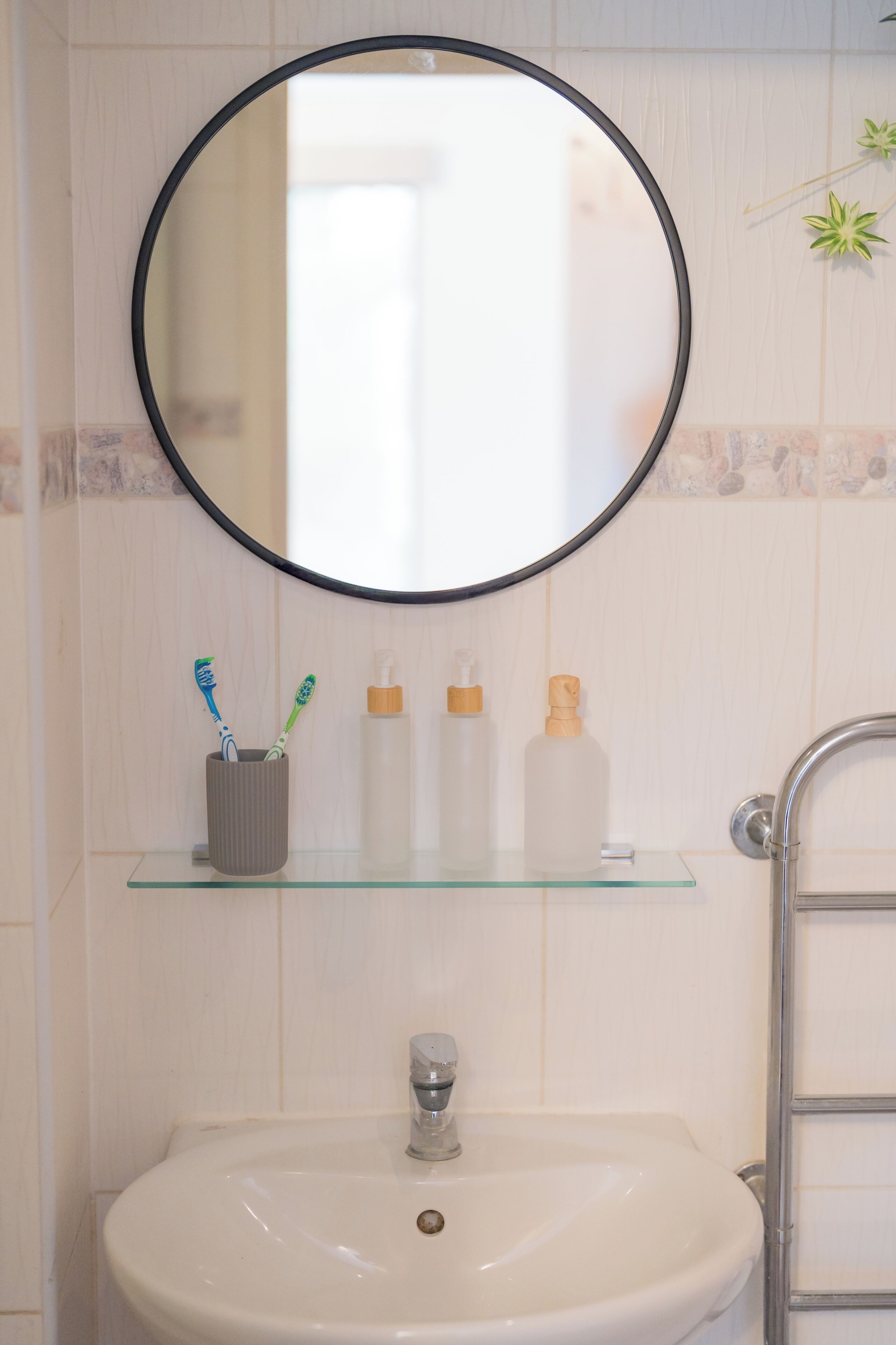 Une salle de bain avec un miroir rond, une étagère en verre où sont posées des brosses à dents et des articles de toilette, et un lavabo avec un robinet.