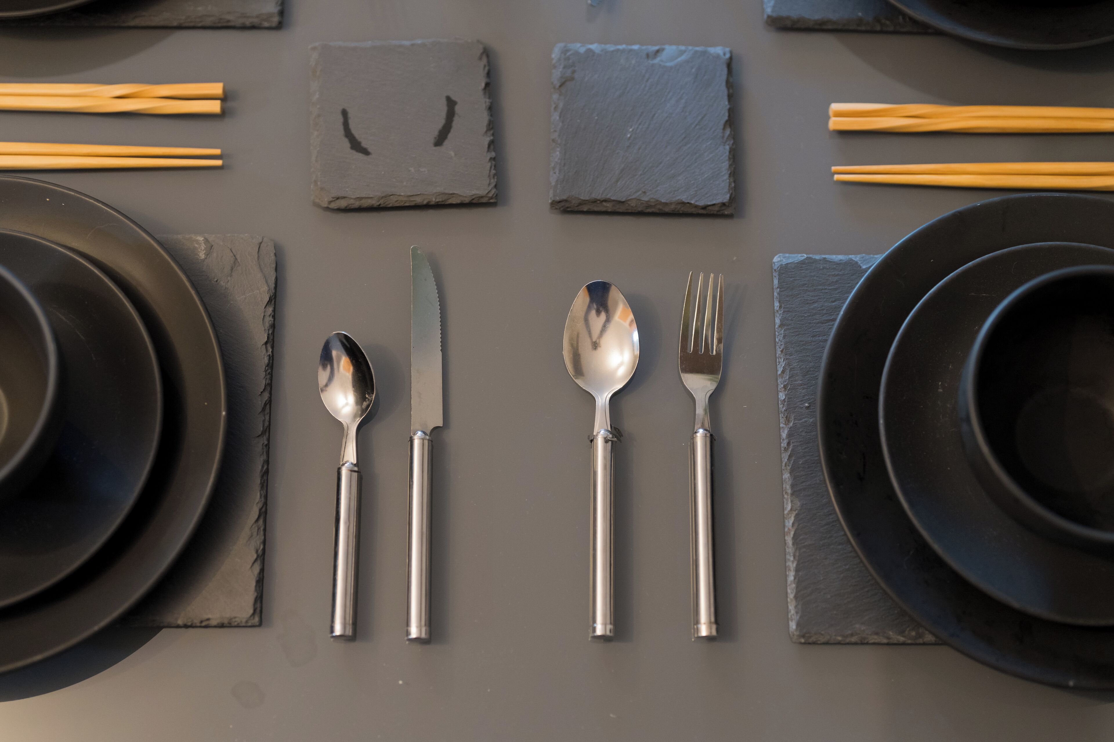 Une table dressée avec des assiettes noires, un couteau, une cuillère et une fourchette, ainsi que des baguettes.