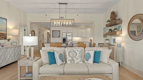 A living room with a sofa, a coffee table, a side table, a chandelier, a kitchen in the background, and a mirror on the wall.