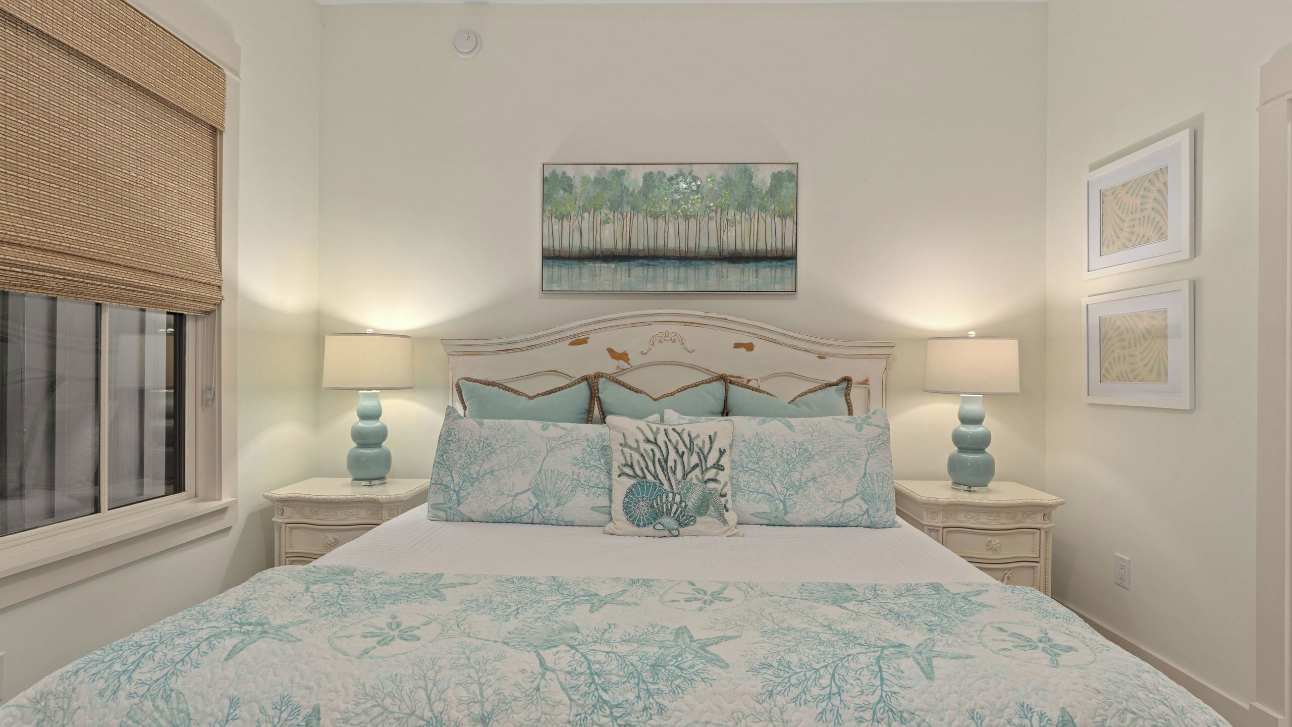 A bedroom with a bed, two nightstands, a window with blinds, a ceiling light, and a painting on the wall.