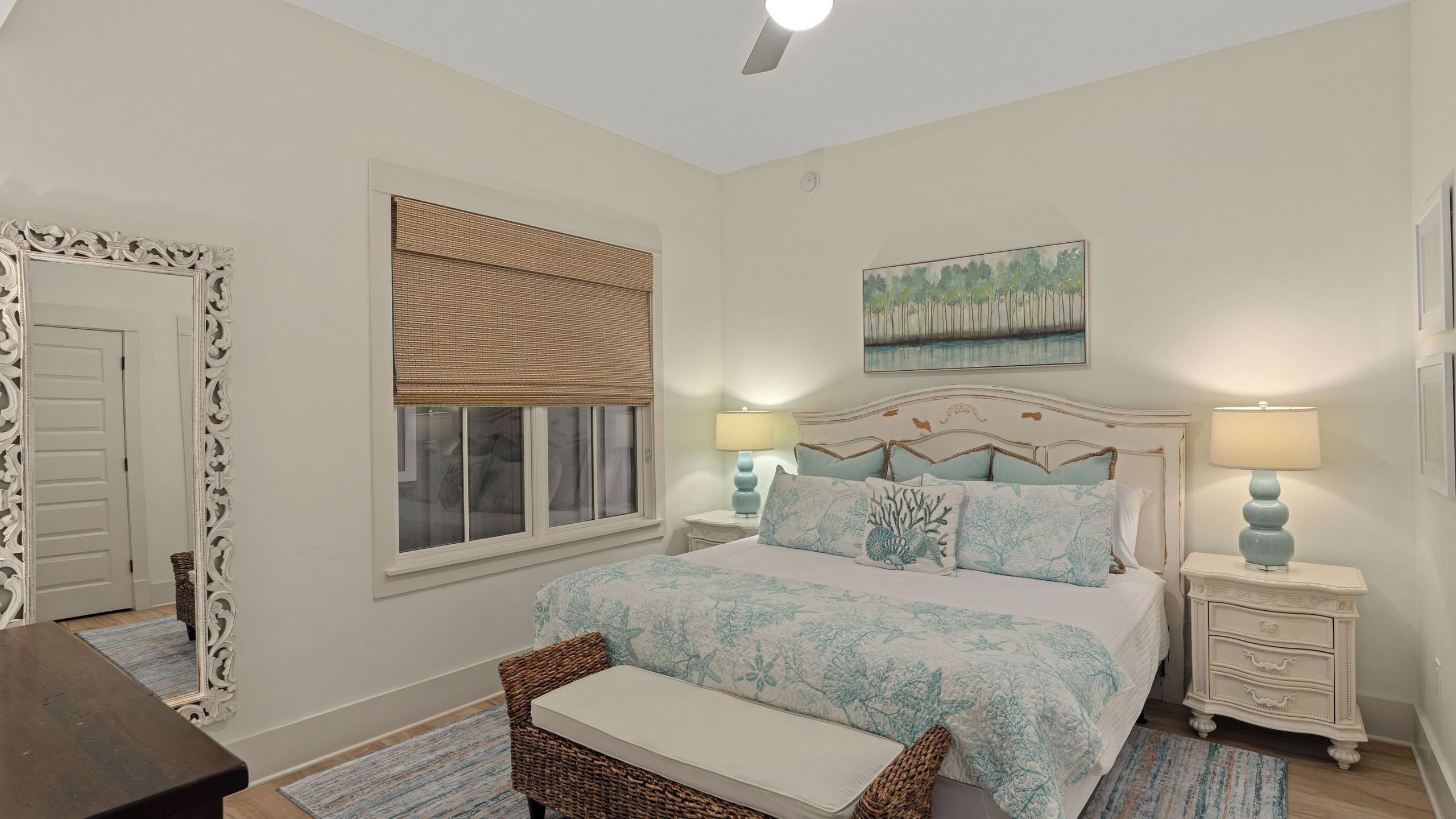 Un dormitorio con cama, dos mesitas de noche, una mesita auxiliar, una silla, ventana con cortina, un cuadro y una puerta.