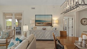 A living room with a sofa, dining table, and chairs, a painting of a beach scene, and a clock on the wall. - 30A Kinda Day by Avantstay Pool + Private Beach Top Floor Redfish (Santa Rosa Beach)