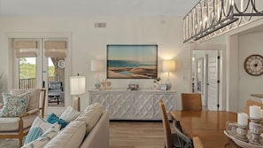 A living room with a sofa, dining table, and chairs, a painting of a beach scene, and a clock on the wall.
