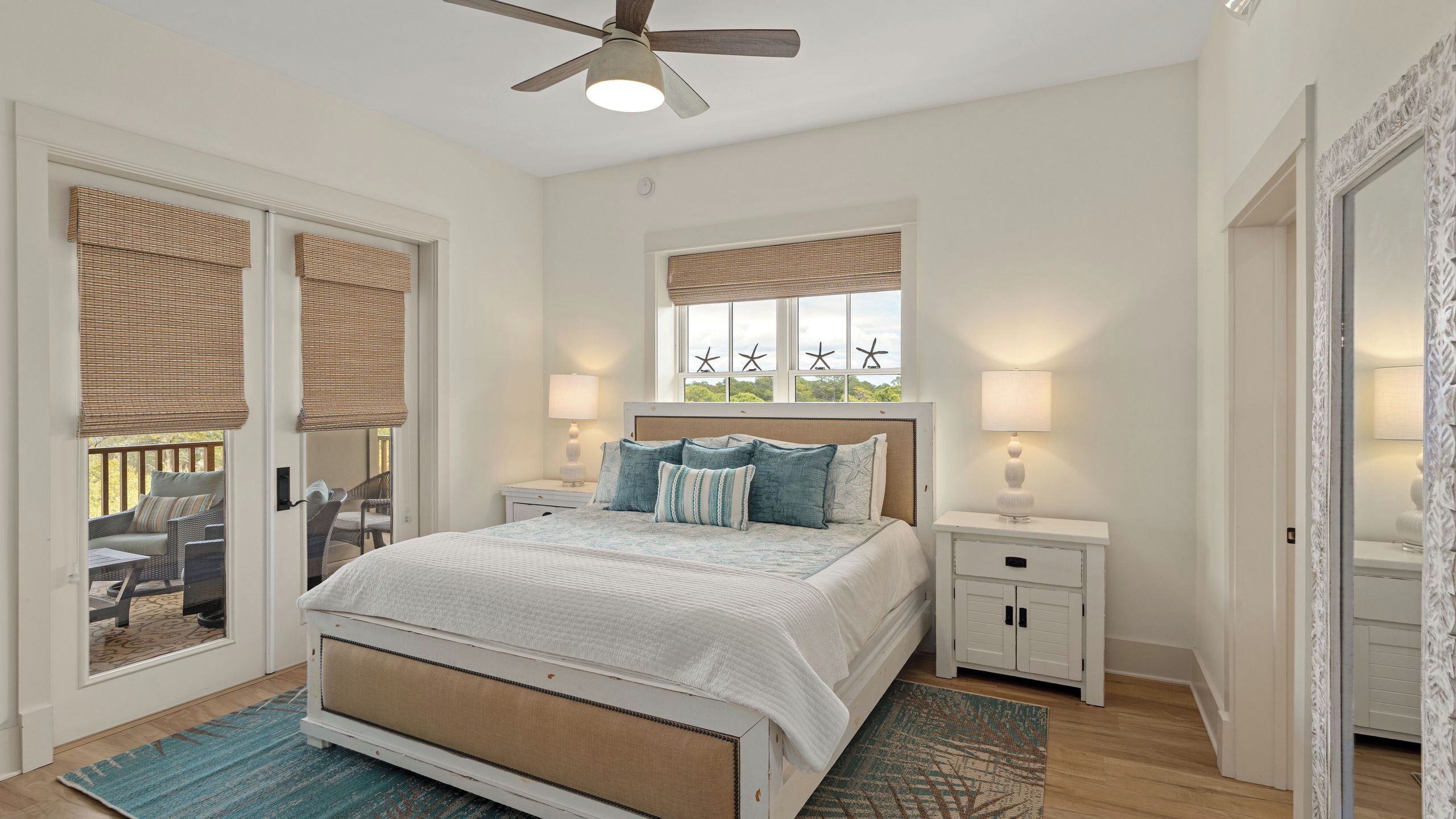 A bedroom with a bed, nightstands, a ceiling fan, a window with starfish decorations, and a balcony with wicker blinds.