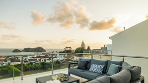 Un balcon avec un canapé gris, une table sur laquelle se trouvent un livre et une théière, et une vue sur la mer et les bâtiments.