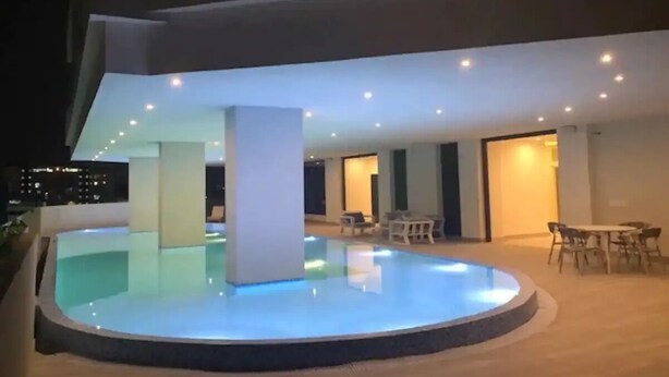 Un área de piscina al aire libre moderna con diseño circular, rodeada por una terraza de madera. Hay varias zonas de estar con mesas y sillas, y el espacio está iluminado por luces empotradas en el techo.