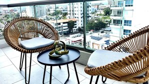 Un balcón con sillas de mimbre y una mesita pequeña, con vista a un paisaje urbano con edificios y cielo despejado.