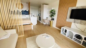 A modern hotel room with a bed, a white sofa, a round coffee table, a TV mounted on the wall, a ceiling fan, and a kitchen area in the background. - Studio Cocoon, sea and marina view. (Marigot)