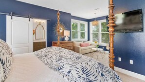 A bedroom with a four-poster bed, a ceiling fan, a television mounted on the wall, and a window with blinds.