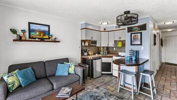Ein kompakter Wohnbereich mit Sofa, Couchtisch und einer kleinen Küchenzeile inklusive Kühlschrank, Mikrowelle und Barhocker.