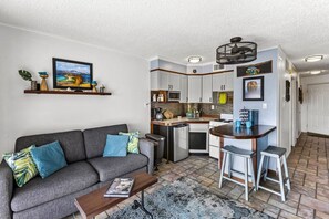 Un espace de vie compact comprenant un canapé, une table basse et un petit coin cuisine équipé d’un réfrigérateur, d’un four à micro-ondes et de quelques tabourets de bar.