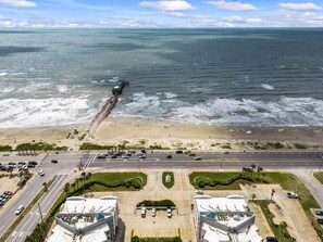 An aerial view of a coastal area with a beach, a pier, and a parking lot with several buildings.