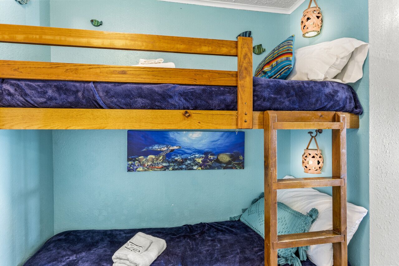 Una cama nido con estructura de madera y una manta azul, una escalera y un cuadro de una escena submarina.