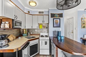 Une cuisine compacte avec des meubles blancs, un four à micro-ondes, une cuisinière et une machine à café. On y trouve un plan de travail en bois et un ventilateur au-dessus.