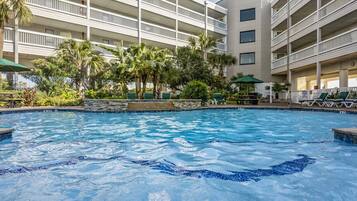 A hotel pool area with a clear view of the pool, surrounded by palm trees and a multi-story building with balconies.