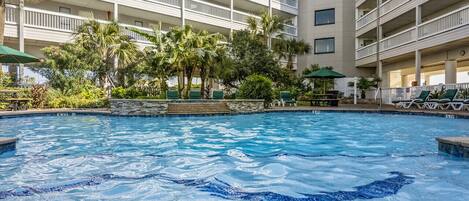 A hotel pool area with a clear view of the pool, surrounded by palm trees and a multi-story building with balconies.