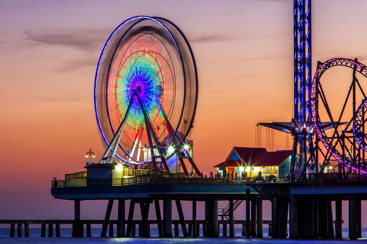 A pier with a large Ferris wheel and a roller coaster, both illuminated at dusk.