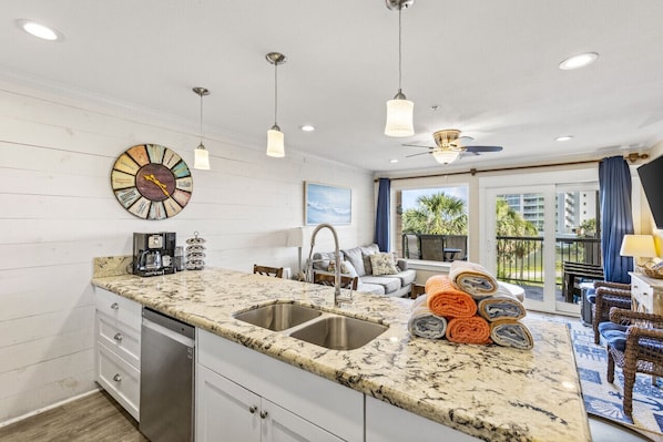 A kitchen with a granite countertop, stainless steel sink, and a coffee maker. There is a dining area with a sofa and a view of palm trees.