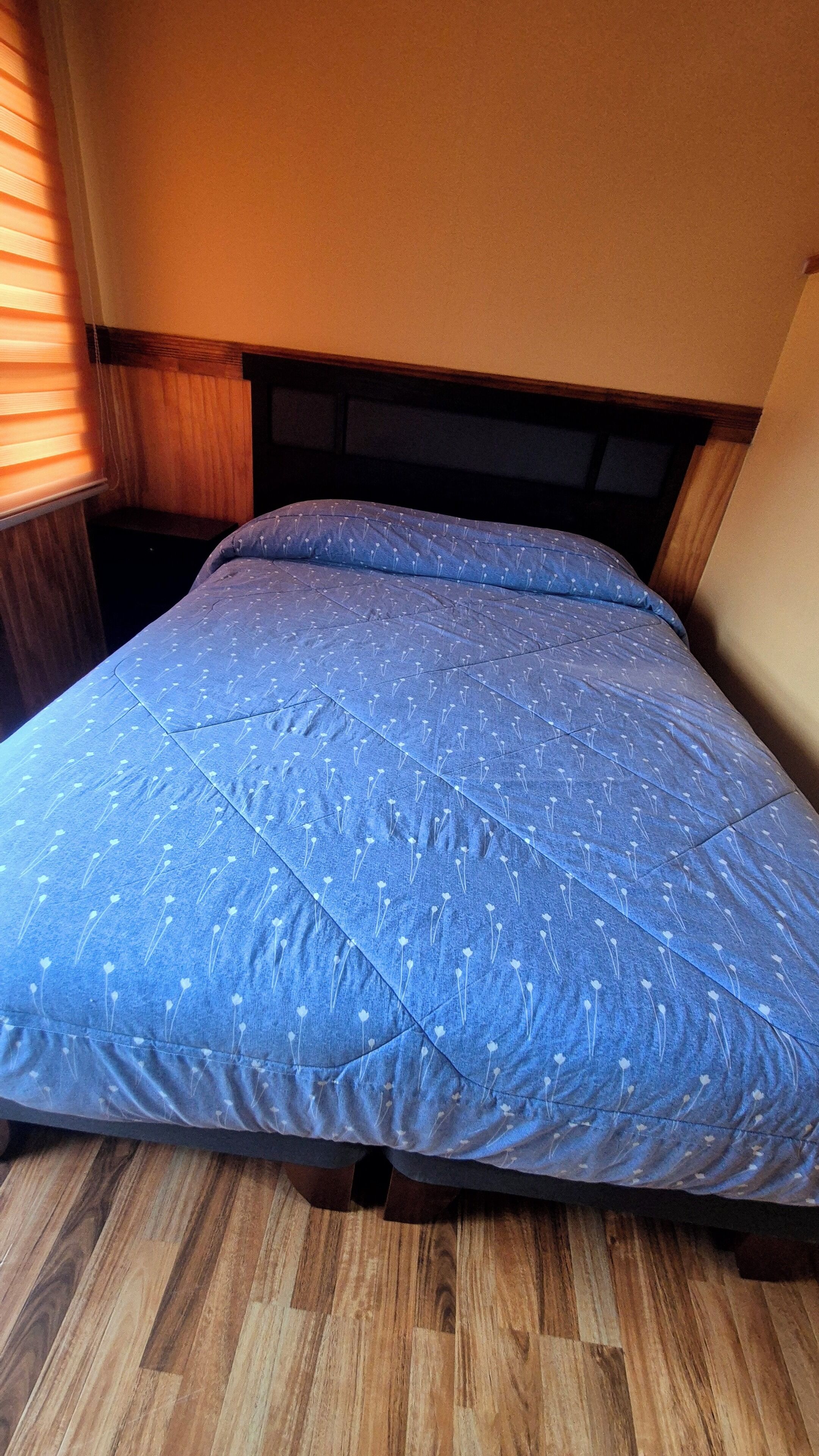 A bed with a blue quilted cover and wooden headboard in a room with wooden flooring and a window with blinds.