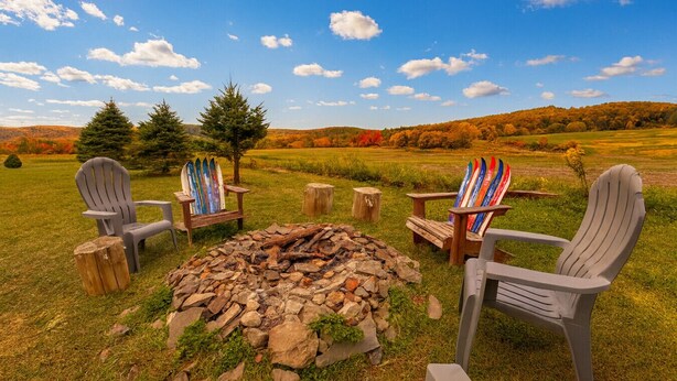 Uma fogueira com quatro cadeiras Adirondack, uma delas com esquis coloridos, situada num campo gramado com árvores e colinas ao fundo.