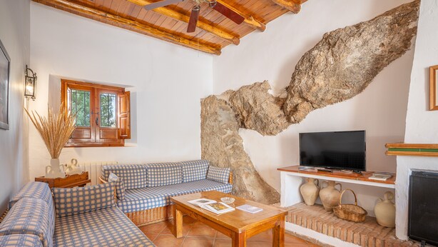 A living room with a checkered sofa, a wooden coffee table, a television, and a stone wall. - Holiday Home 'Casa Rural La Roca' with Mountain Views, Shared Pool and Wi-Fi (La Zubia)