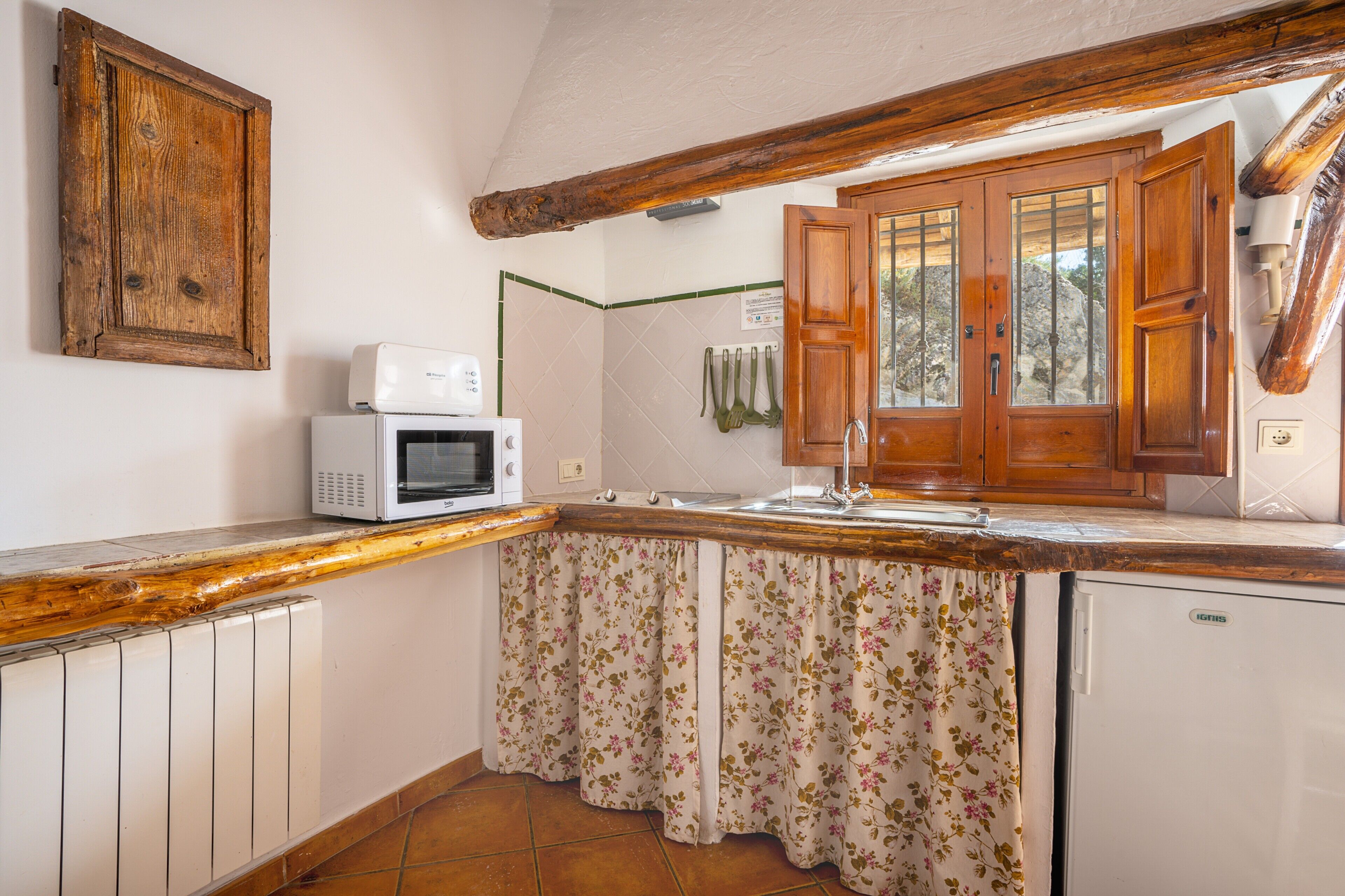 A kitchen with a microwave, a refrigerator, wooden cabinets, a window with wooden shutters, and a radiator.