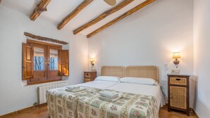 A bedroom with a bed, wooden beams, a window with shutters, two bedside tables with lamps, and a radiator. - Holiday Home 'Casa Rural La Roca' with Mountain Views, Shared Pool and Wi-Fi (La Zubia)