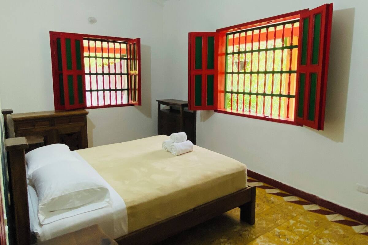 A hotel room with a bed, two windows with red frames, a small table, and a chair.