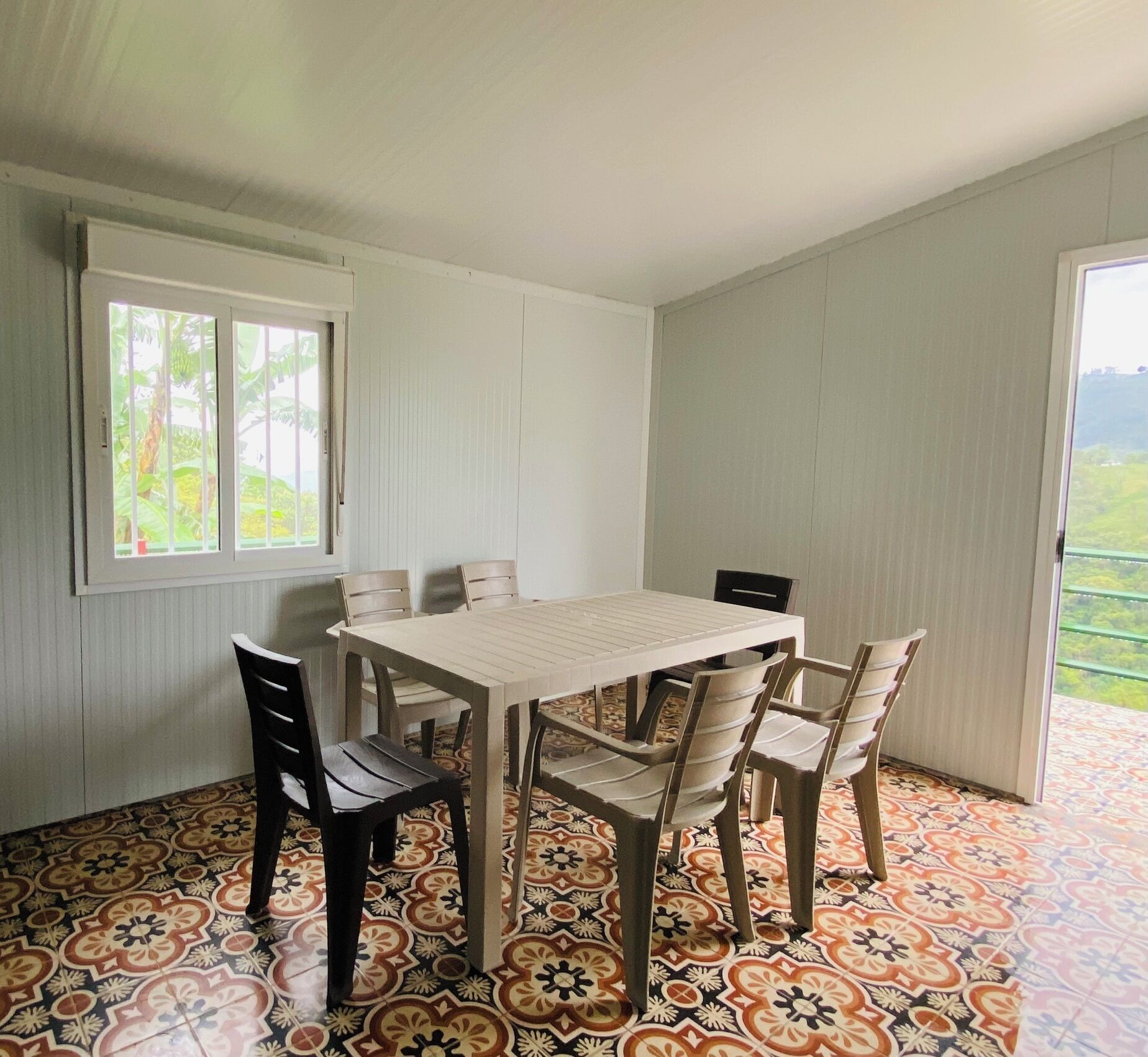 A dining area with a table and chairs, a window with a view of greenery, and patterned floor tiles.