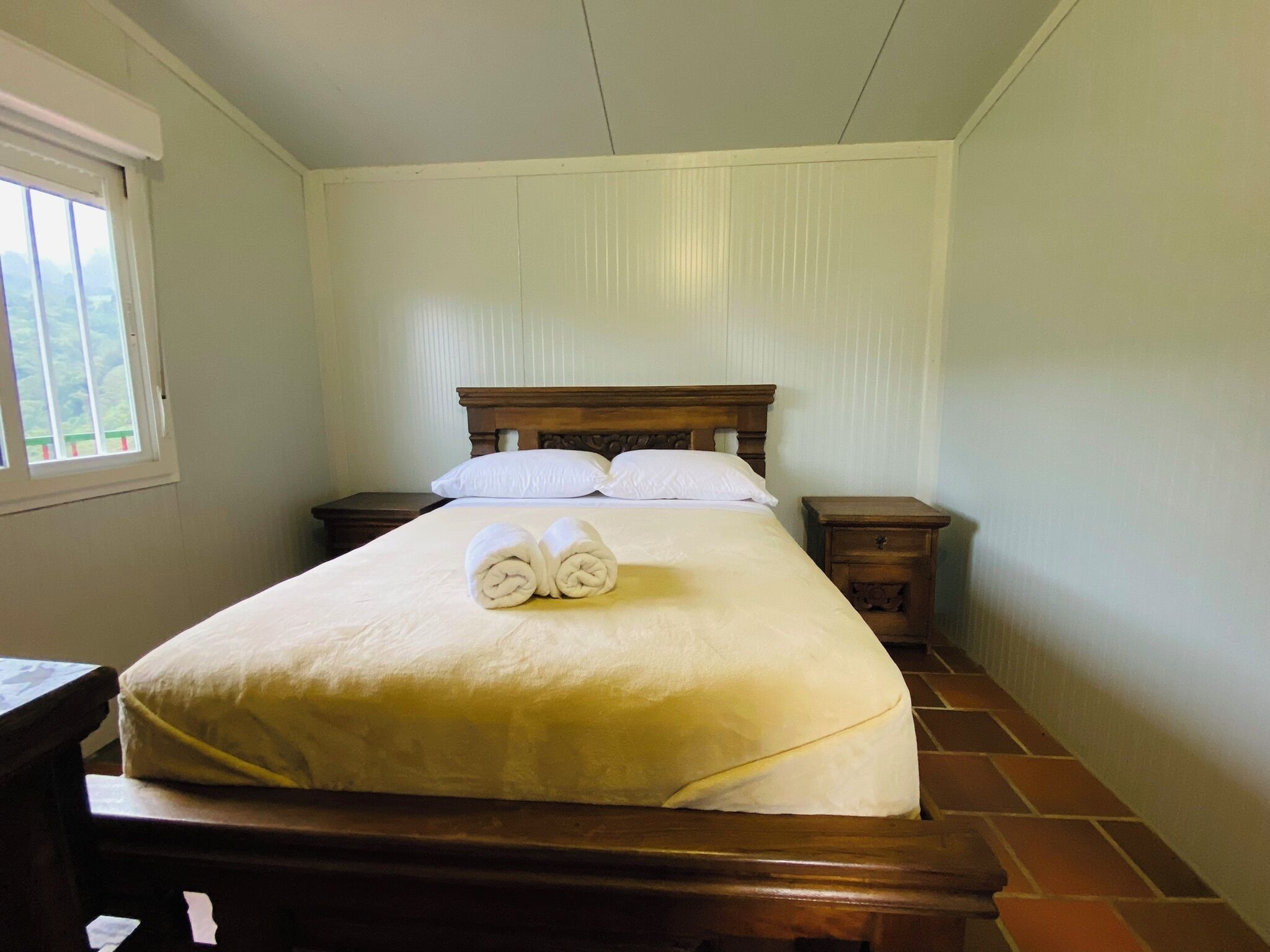 Una cama bien hecha con cabecera de madera, dos mesitas de noche y una ventana con vistas a zonas verdes.