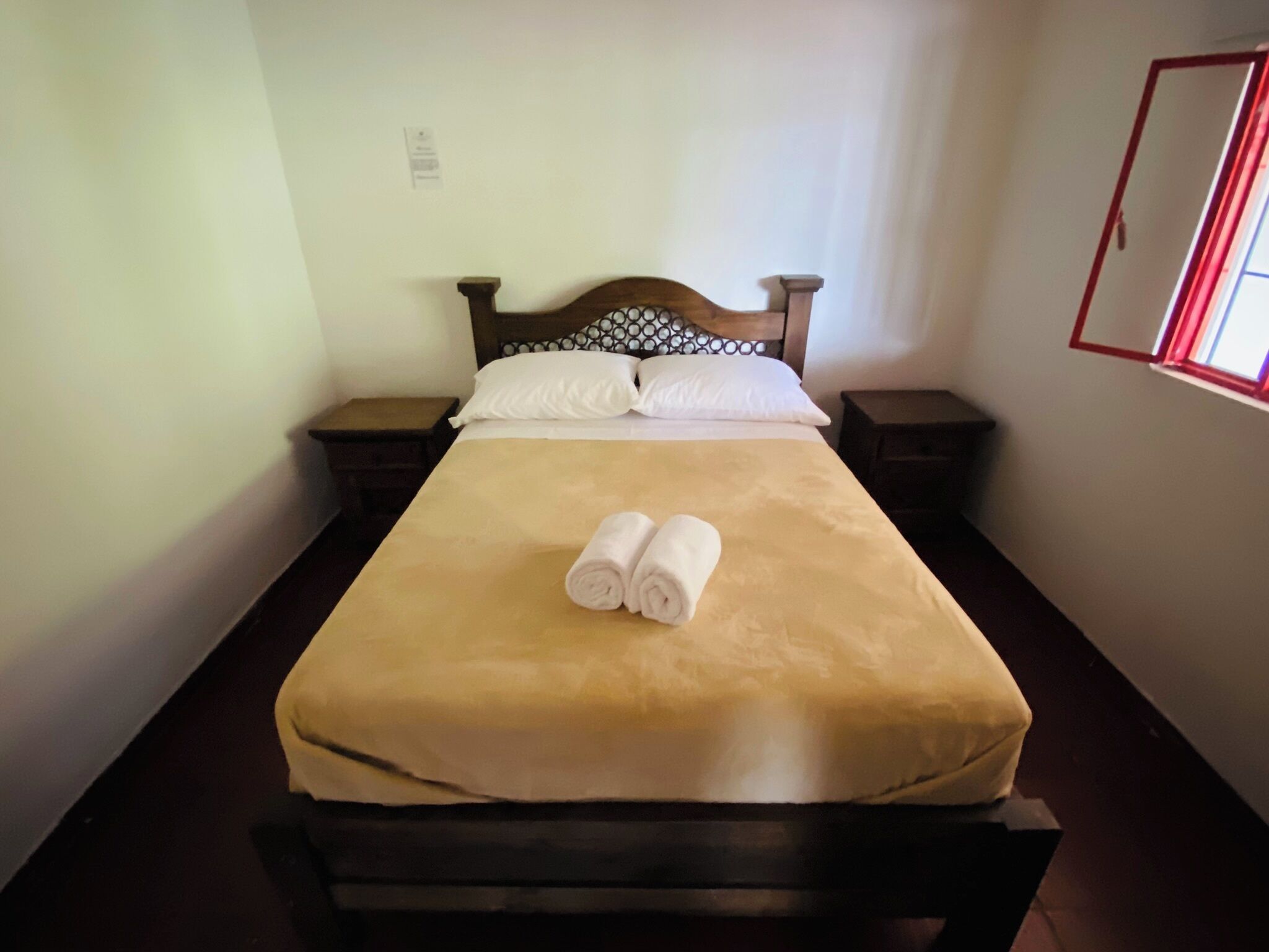 Un lit bien fait, recouvert d’une couverture marron, avec deux serviettes enroulées, une tête de lit en bois, deux tables de chevet et une fenêtre à cadre rouge.