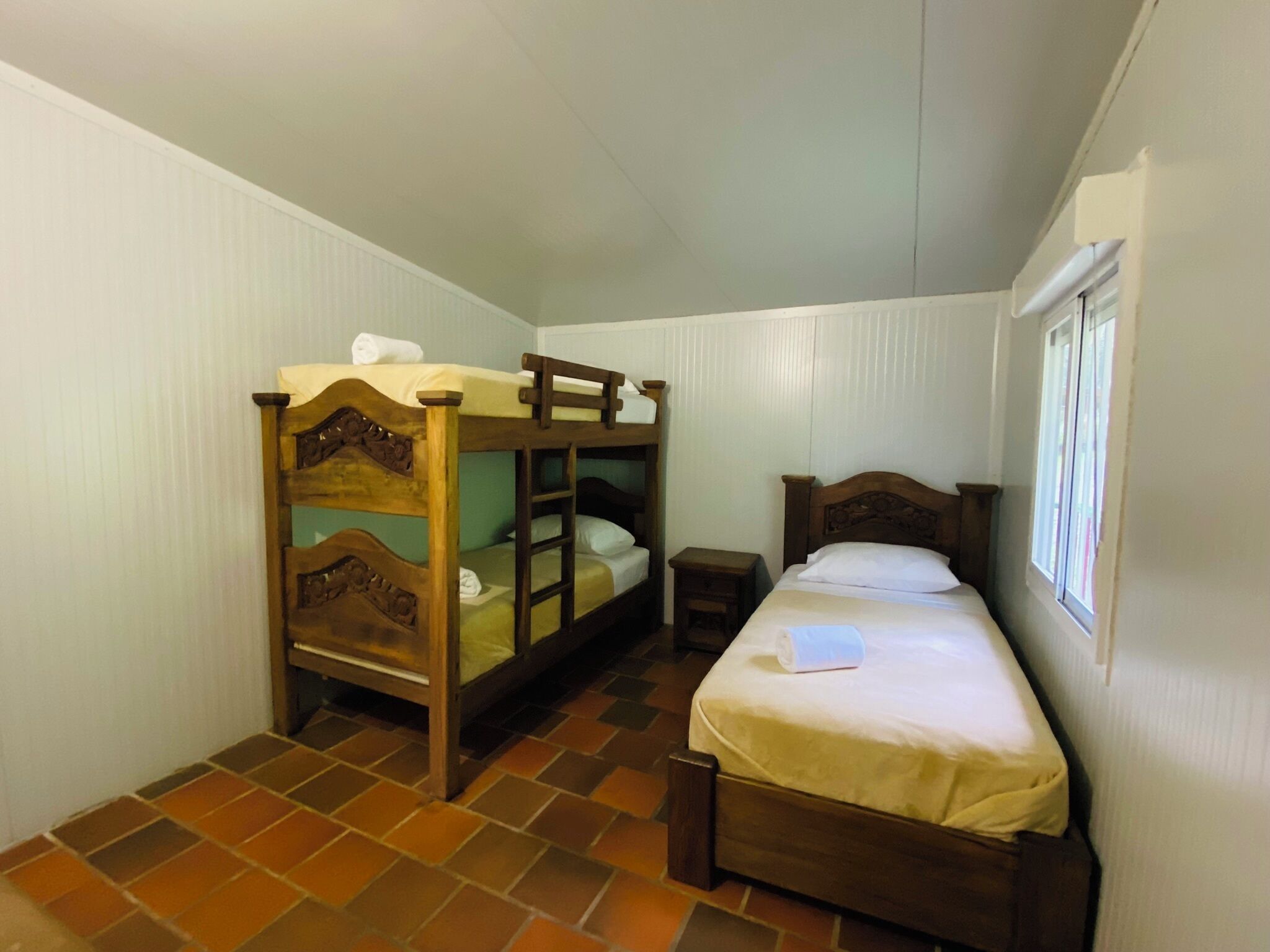 A room with two wooden bunk beds, a single bed, a small bedside table, and a window.