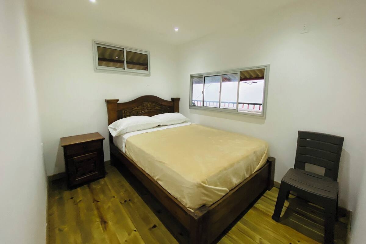 A bedroom with a wooden bed, a nightstand, a chair, a window with a view, and a ceiling light.