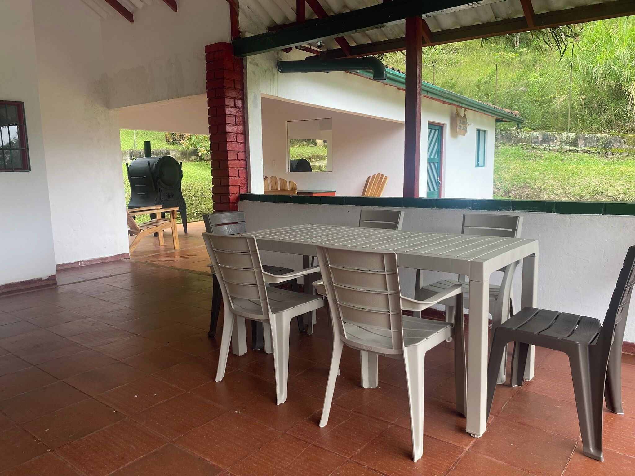 A covered outdoor dining area with a table and chairs, a grill, and a view of greenery.
