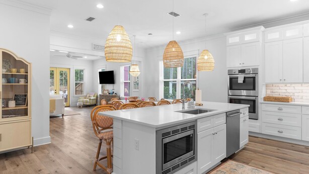 A modern kitchen with white cabinetry, a central island with a sink, and a built-in oven. There are wicker pendant lights and a large window providing natural light. - Priv Heated Pool + Hot Tub, Boat-friendly, Sleeps 16! Good Times by Avantstay (Santa Rosa Beach)
