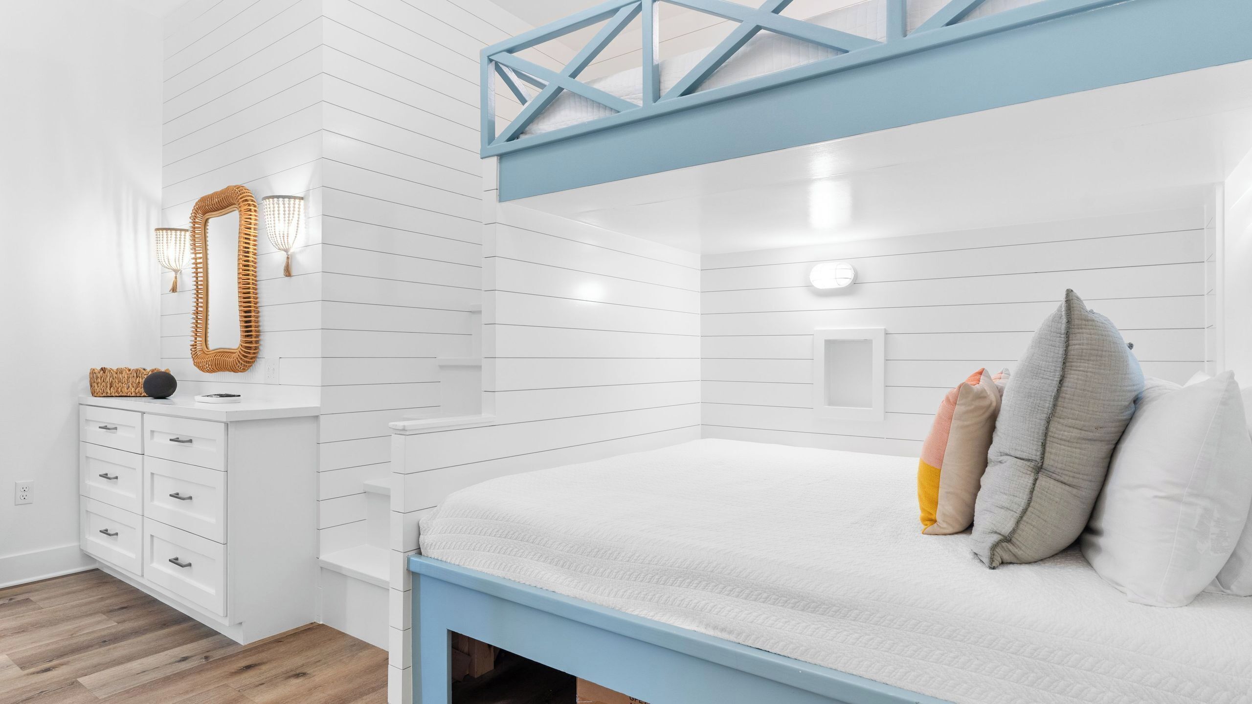 A bedroom with a bunk bed, white walls with horizontal lines, a dresser with drawers, a mirror, and a bedside table with a lamp.