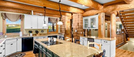Kitchen - Large island with a cooktop, pendant lights, and bar seating under massive log beam ceilings with white and green cabinetry.