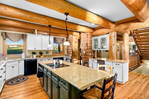 Kitchen - Large island with a cooktop, pendant lights, and bar seating under massive log beam ceilings with white and green cabinetry.