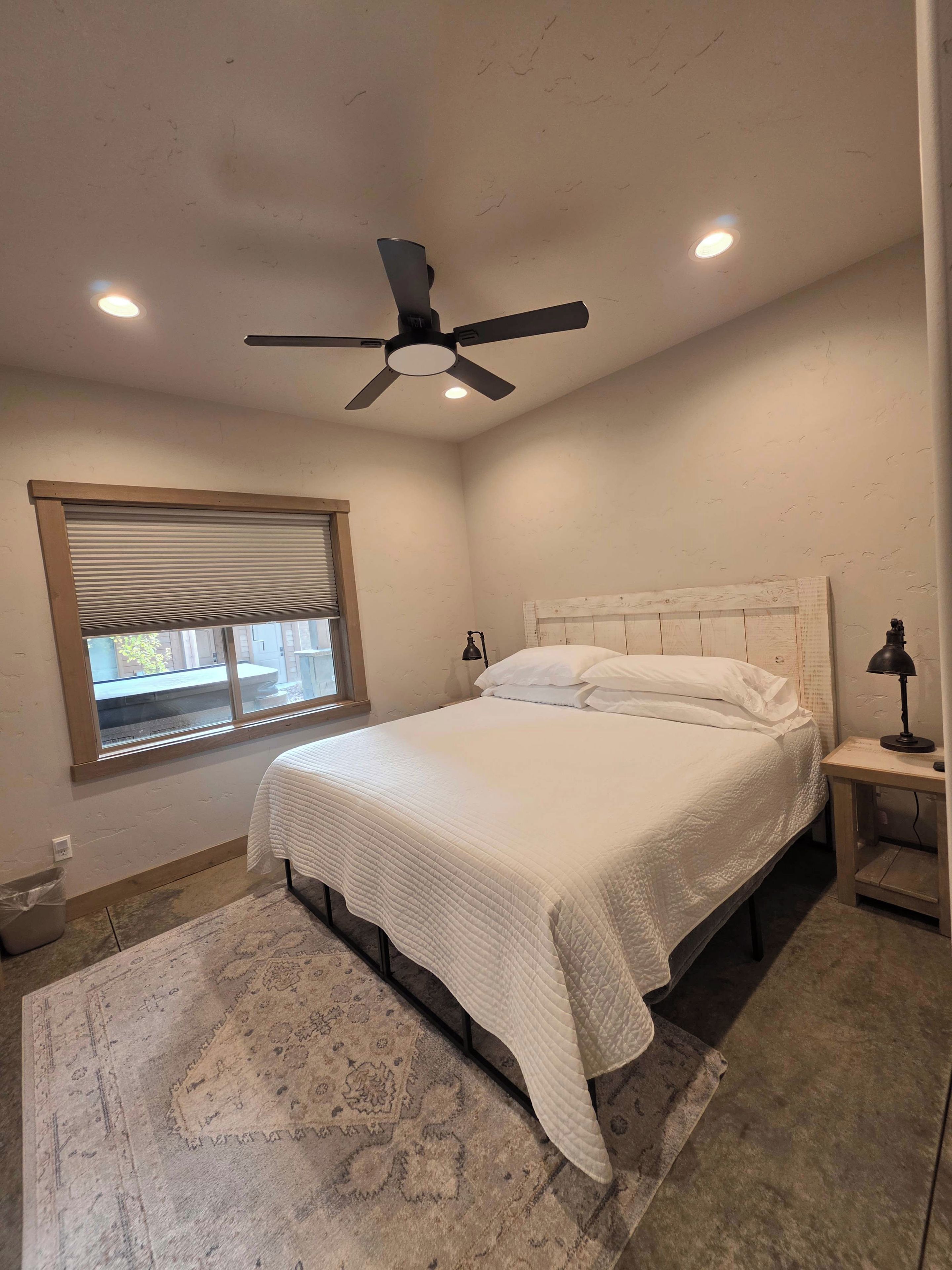 Un dormitorio con cama, ventilador de techo, ventana y mesita de noche.