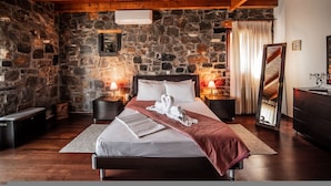 A bedroom with a stone wall, wooden ceiling beams, a bed with white linens, a red throw blanket, and a mirror on a dresser. - Villa Gaia With Private Pool (Agios Nikolaos)