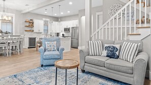 Ein modernes Wohnzimmer mit einem grauen Sofa, einem blauen Sessel und einem Couchtisch aus Holz. Im Hintergrund ist eine Treppe mit weißen Geländern und Holzstufen zu sehen.