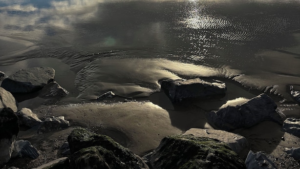 A beach with calm waters, rocks, and a cloudy sky with the sun setting or rising.