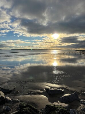 A beach with calm waters, rocks, and a cloudy sky with the sun setting or rising.