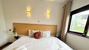 A neatly made bed with white linens and rolled towels, a wooden headboard, two wall-mounted lights, and a window with curtains.