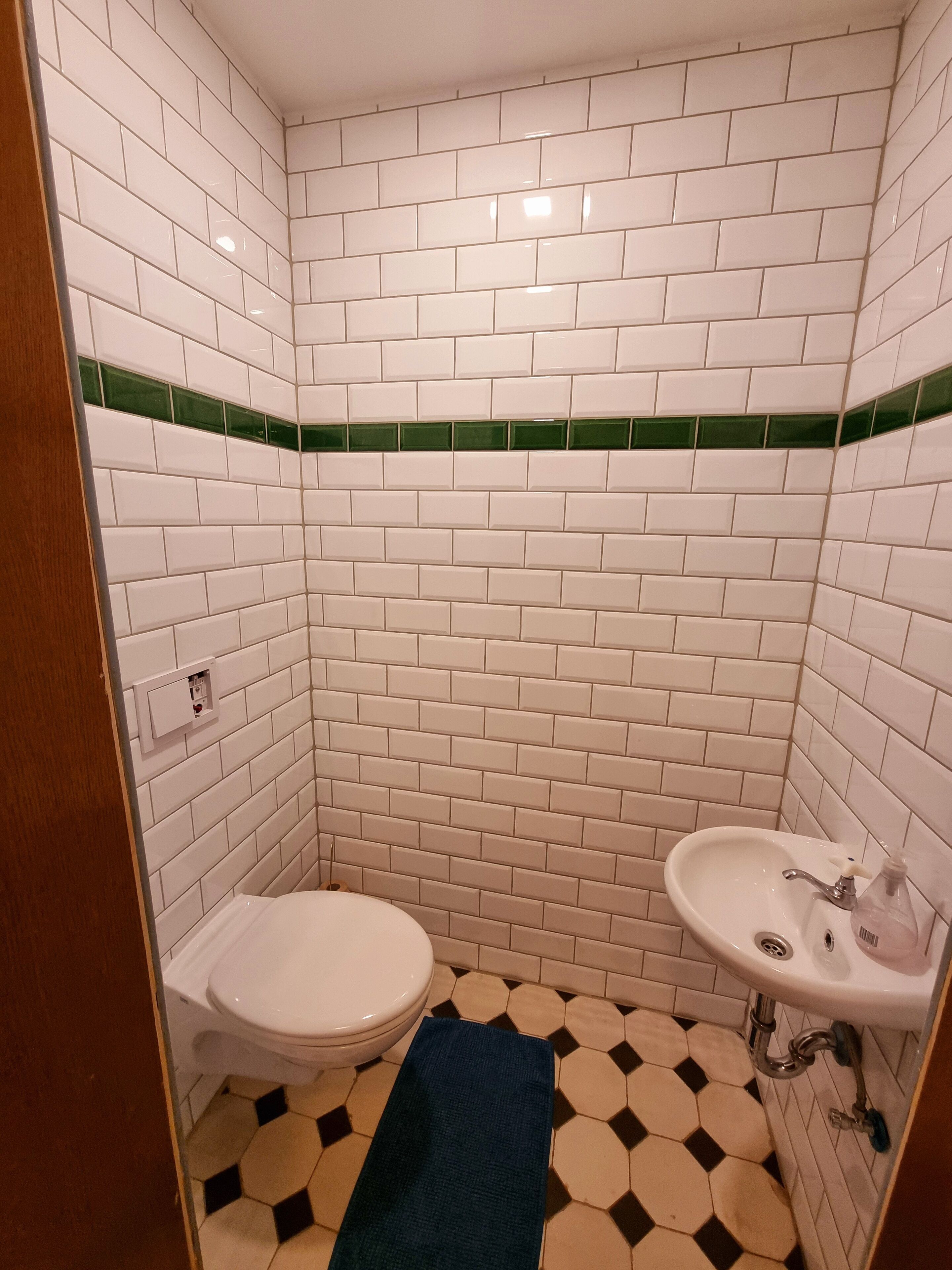 A small bathroom with white subway tiles, a green accent strip, a toilet, a sink, and a blue towel on the floor.